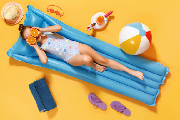 Little girl in a swimsuit and half glasses of orange resting on an inflatable blue mattress on a yellow background. Little girl in a swimsuit and half glasses of orange resting on an inflatable blue mattress on a yellow backgroun. little girls in bathing suits stock pictures, royalty-free photos & images