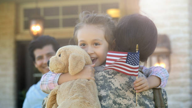 Little girl is lifted into hug by a young adult female in uniform A woman returning home in uniform, lifts a smiling child and her stuffed toy into the air for a hug, while a smiling man watches. soldiers returning home stock pictures, royalty-free photos & images