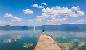 Small child (girl) standing on a wooden bridge, at the edge of the lake, looking at a sailboat (yawn). There are mountains in the background and clouds reflecting in the water. The day is sunny and the atmosphere is very peaceful.