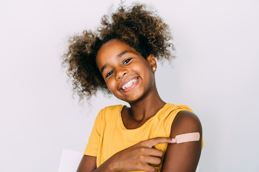 Cute smiling girl pointing at her arm with a bandage after receiving COVID-19 vaccine. Shot of little african-american girl showing her shoulder after getting coronavirus vaccine.