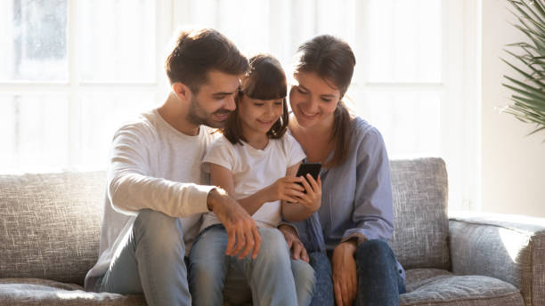 Little girl showing to parents favourite game play online Little daughter sitting with parents holding smart phone showing playing games using online apps, mom dad having bought new device teaching kid, people having video call. Modern wireless tech concept free adult webcams stock pictures, royalty-free photos & images