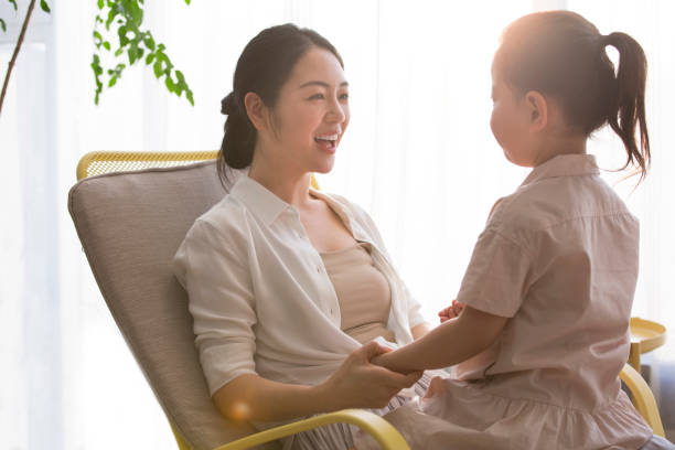 A little girl sits on her young mother's lap as they rock the rocking chair and sing children's songs and tell fairy tales - stock photo Suitable for parenting, family education, early childhood development, child care, family decoration, intellectual development, hands-on, children's clothing, expatriate Immigration, preschool education, print advertising. Portrait. chinese girl hairstyle stock pictures, royalty-free photos & images