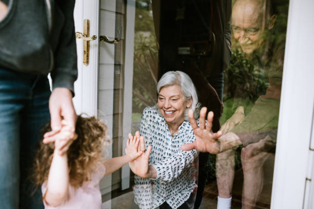 Little Girl Visits Grandparents Through Window A mother stands with her daughter, visiting senior parents but observing social distancing with a glass door between them.  The granddaughter puts her hand up to the glass, the grandfather and grandmother doing the same.  A small connection in a time of separation during the Covid-19 pandemic. quarantine stock pictures, royalty-free photos & images