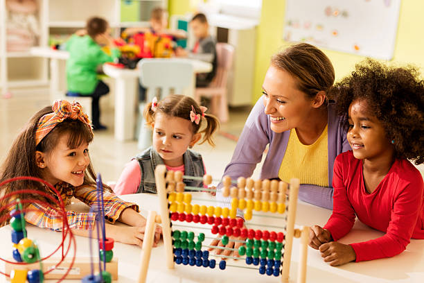 Little girls and teacher learning at preschool. Smiling teacher and little girls talking and playing with educational toys at preschool. Other children are in the background. abacus stock pictures, royalty-free photos & images