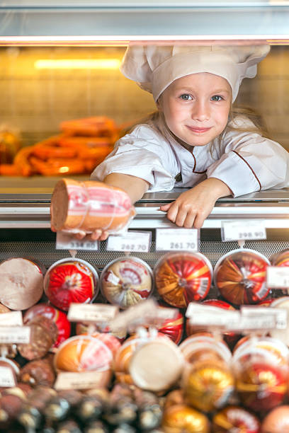 Little salesgirl sausage Cute little girl in a seller uniform holding out her hand with a sausage standing behind the counter with sausage salesgirl stock pictures, royalty-free photos & images