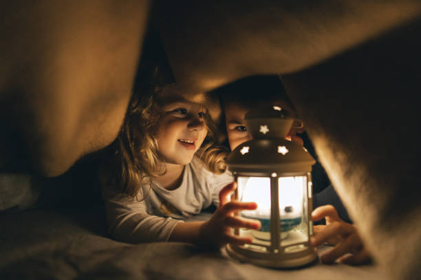 Little sister and her brother lying on bed under blanket in dark room and holding rustic lantern lamp stock photo