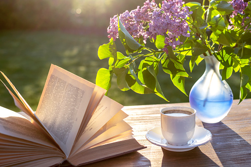 Little white cup of espresso coffee, opened book, blue semi-transparent vase with purple lilac flowers on rustic wooden table in the garden at spring morning after sunrise or at evening before sunset