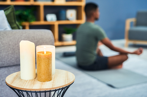 Shot of candles lit in a living room with a young man meditating in the background