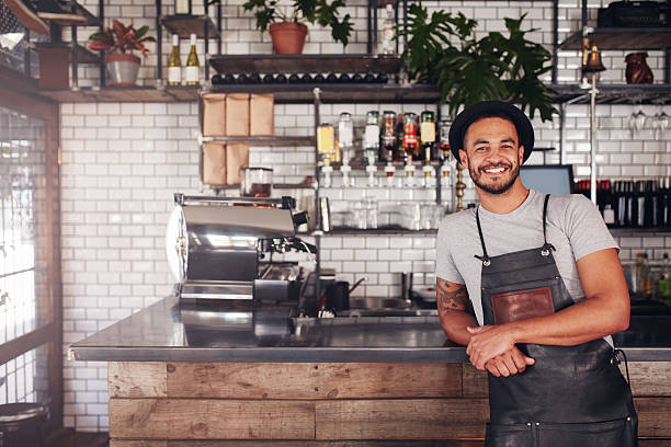 Local coffee shop owner Portrait of young man standing at the counter in his cafe. Coffee shop working in apron and hat smiling at camera. barista stock pictures, royalty-free photos & images