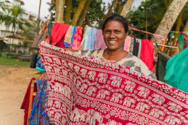 Local Sri Lankan Indian woman senior positive smiling cheerful woman, the small business owner, demonstrating textile in her open-air clothing and souvenirs store on the beach. Local Sri Lankan Indian woman, the small business owner, demonstrating textile in her open-air clothing and souvenirs store on the beach. sri lanka women stock pictures, royalty-free photos & images