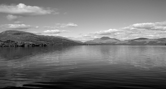 Wide angle, panoramic view of the pristine waters of Loch Lomond with the mountains of the Scottish Highlands in the background. The clouds above reflect in the calm waters of the loch. Loch Lomond is the largest loch/lake in Great Britain by surface area.