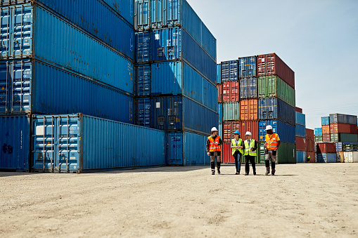 Mid distance view of diverse cargo handlers and supervisors approaching camera in reflective vests and hardhats amidst stacks of cargo containers.