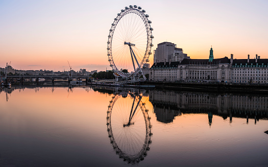 The iconic ring of the London Eye Ferris wheel reflecting in the tranquil waters of the River Thames at sunrise, Westminster, UK.