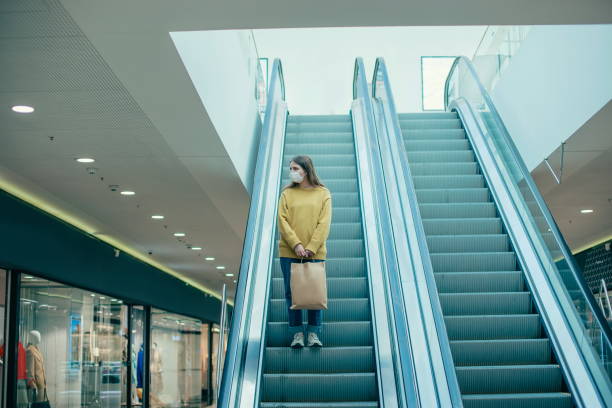 lone woman in a protective mask standing on the escalator steps lone woman in a protective mask standing on the escalator steps . photo with a copy-space shopping mall stock pictures, royalty-free photos & images