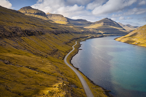 Lonely empty country road along Funningsfjordur Fjord in late summer, Aerial Drone Point of view along the rural national road on Funningsfjørður on Eysturoy Island in between green hills and North Atlantic Ocean Fjord Inlet. Funningsfjørður Fjord, Eysturoy Island, Faroe Islands, Kingdom of Denmark, Nordic Countries, Scandinavia, Europe.