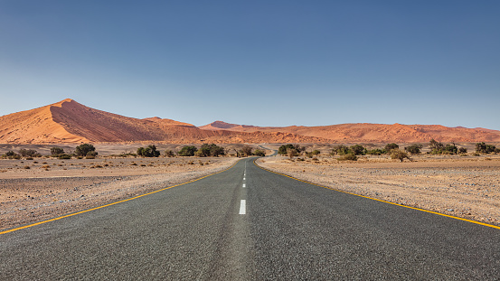 Empty Open Desert Road inside the arid vast Desert Landscape of the Sossusvlei Nambib Desert of Namibia. Giant Sand Dunes roadside the Highway. Panorama Shot under blue summer skyscape. Namibian Desert, Sossusvlei, Namibia, Southern Africa.