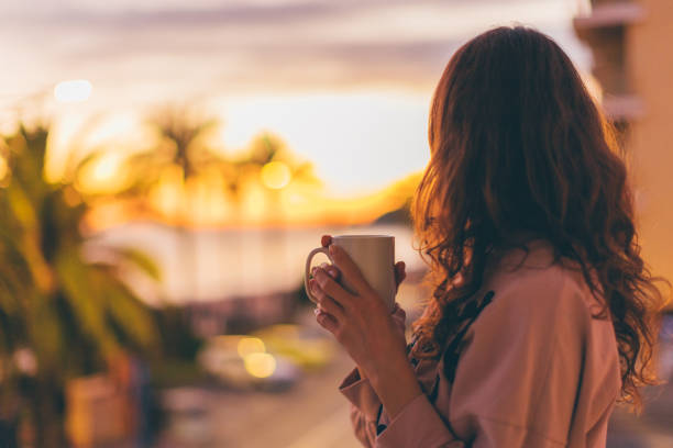Lonely romantic girl drinking coffee looking at sunset. Rear view of a girl with cup of hot drink balcony stock pictures, royalty-free photos & images