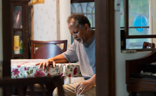 Lonely senior man sitting at home, depressed An African-American senior man in his 60s sitting at home at the kitchen table. He has a serious, sad expression on his face, looking down toward the floor. He is lonely and depressed sad old black man stock pictures, royalty-free photos & images