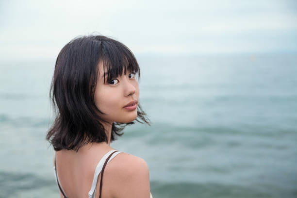 Lonly Young Woman By The Sea A lonely Japanese woman is looking at the camera over her shoulder by the sea. japanese girl stock pictures, royalty-free photos & images