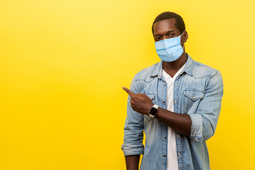 Look, advertise here! Portrait of positive man with surgical medical mask pointing left side and smiling at camera, showing empty space for advertise. indoor studio shot isolated on yellow background