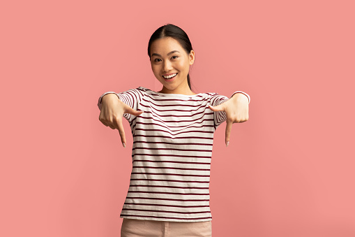 Look At This. Positive Asian Female Pointing Down With Two Hands, Cheerful Korean Woman Demonstrating Free Copy Space For Your Design And Advertisement While Standing Isolated On Pink Background