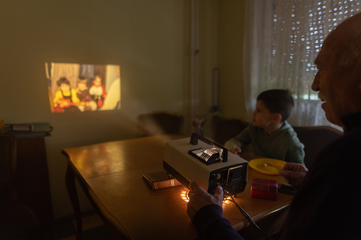 Grandfather and grandson sitting at the table and looking photos on vintage slide projector at elderly man's home.