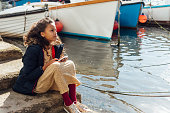 A young girl sitting on steps at a commercial dock while on vacation in Polperro, Cornwall. She is holding a mobile phone while looking off into the distance.