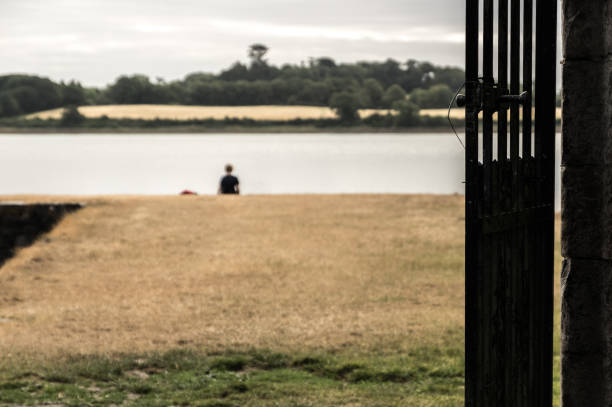 Looking through a gateway towards a boy sitting on an abandoned pier Looking through a gateway, past a black iron gate, towards a teenage boy (out of focus) sitting on an abandoned pier or jetty.  Strangford, County Down, Northern Ireland. strangford lough stock pictures, royalty-free photos & images