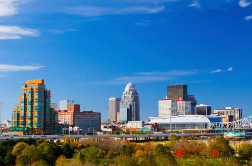 The skyline of downtown Louisville, Kentucky featuring the skyscrapers of the city against a bright blue sky. White clouds can be seen around the edges of the photo. The trees in the lower foreground are green, with some beginning to turn orange and red for the autumn season.