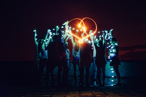 Couple of friends having a party on the rooftop of the building at night, making a heart shaped light effect and holding christmas lights 