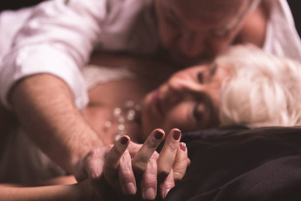 Love kissing your body Elder couple lying together on a bed in an erotic love hug with intertwined fingers sexy old man stock pictures, royalty-free photos & images