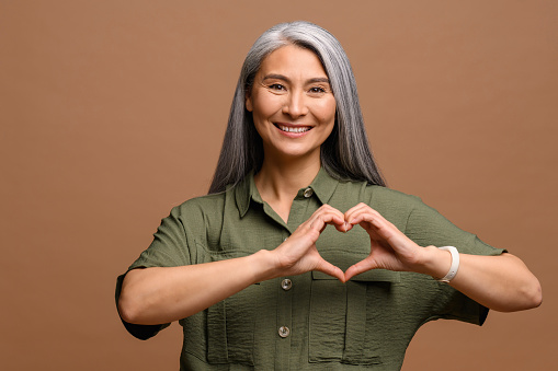 Love you. Portrait of attractive romantic mature woman standing and making heart with hands, while smiling playfully. Indoor studio shot isolated on brown background