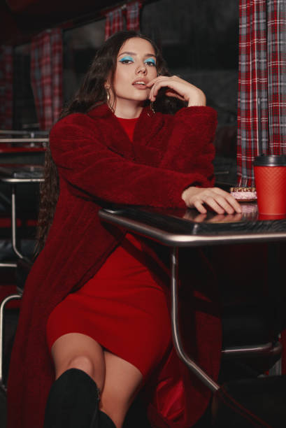 Lovely girl sitting in cafe bus (bistro). Retro (vintage) portrait of beautiful stylish young woman in restaurant, wearing red dress and fur coat stock photo