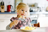 Lovely toddler girl eating healthy fried potatoes for lunch. Cute happy baby child in colorful clothes sitting in kitchen of home, daycare or nursery. Kid eats vegetables