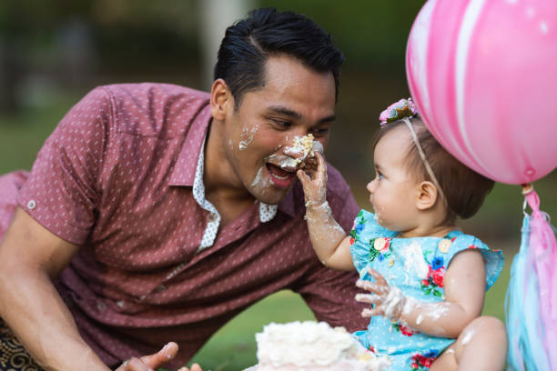 Loving dad celebrating his babys first birthday with cake Adorable mixed race baby girl smears a handful of cake and frosting on her dads face while celebrating her first birthday outside. The family is social distancing and staying at home due to the Covid-19 pandemic and are marking this birthday from the safety of their homes backyard. Milestones, new normal and childhood concepts. baby playing mess stock pictures, royalty-free photos & images