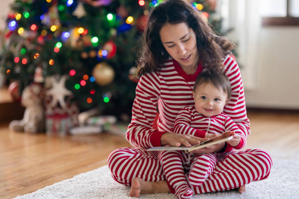 Loving mom reading the Christmas story to her baby An adorable mixed race baby sits in her mother's lap and smiles as they read a book together. The family is celebrating the baby's first Christmas with a cozy day at home. christmas story telling stock pictures, royalty-free photos & images