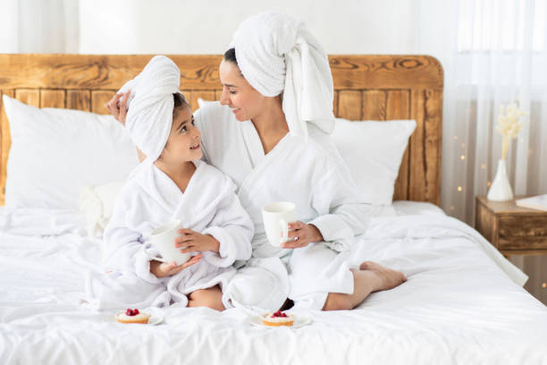Loving mother enjoying tea and cake with little daughter Loving young mother enjoying tea and cake with little daughter after spa procedures at hotel. Mom and female kid with heads wrapped in towels wearing bathrobes, sitting on bed at home, copy space hot middle eastern girls stock pictures, royalty-free photos & images