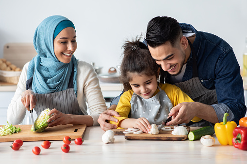Loving Muslim Parents Teaching Their Cute Little Daughter How To Cook Healthy Vegetarian Food, Islamic Family Of Three Having Fun In Kitchen Together, Enjoying Preparing Dinner At Home, Closeup Shot