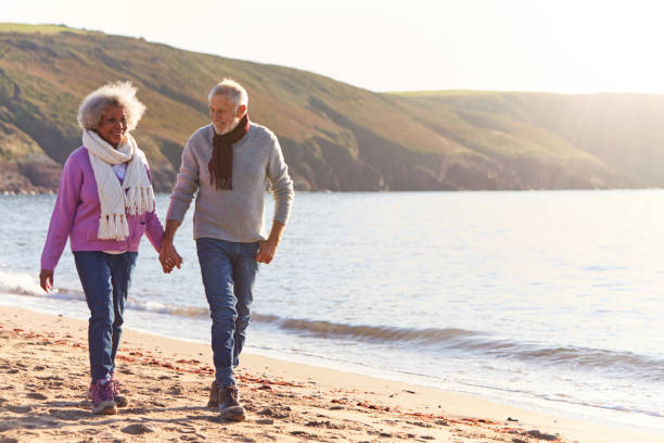 loving retired couple holding hands as they walk along shoreline on winter beach vacation - aktiver senior stock-fotos und bilder