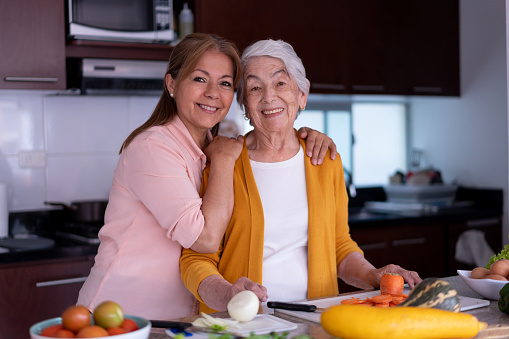 Loving senior mom and daughter preparing a meal together facing camera with a toothy smile at home - Lifestyles