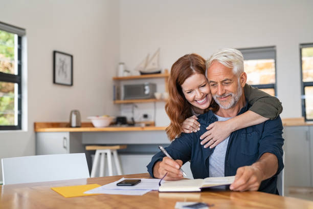 Loving woman embracing senior man Loving mature wife embracing husband from behind while writing in book. Happy middle aged couple making to do list of purchases and discussing future plans. Cheerful senior man working at home on wooden table with beautiful woman hugging him from behind, copy space. mature couple stock pictures, royalty-free photos & images