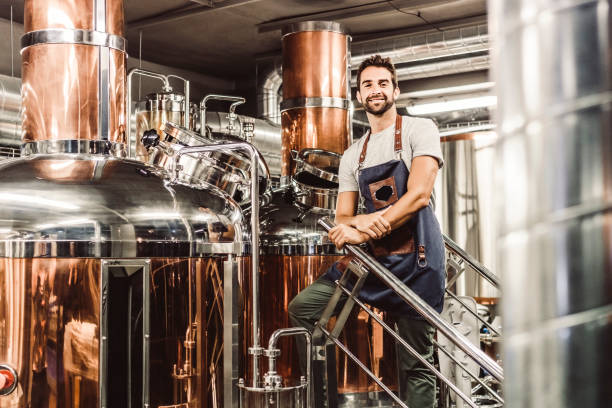 Low angle portrait of male manager at brewery Low angle view of manager standing by distillery stills. Portrait of handsome male owner is on staircase at brewery. He is wearing apron. brewery stock pictures, royalty-free photos & images
