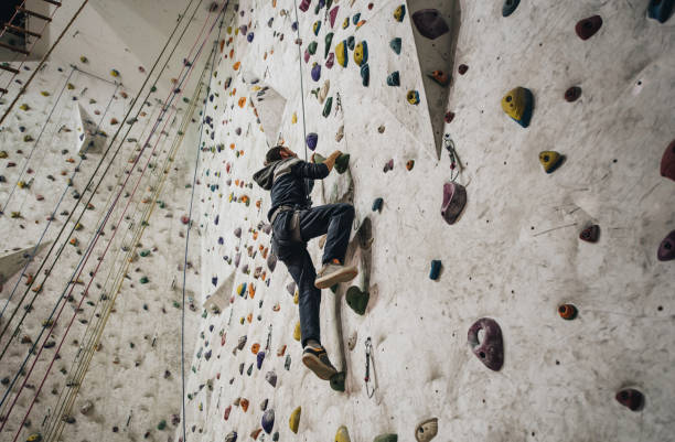 Low angle view of athletic man climbing on the wall in a gym. Below view of a full length male athlete exercising wall climbing. bouldering stock pictures, royalty-free photos & images