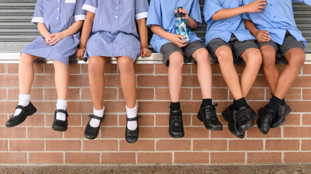 Low section view of five school children sitting on brick wall wearing school uniform Children's legs and feet in black shoes hanging down elementary student stock pictures, royalty-free photos & images