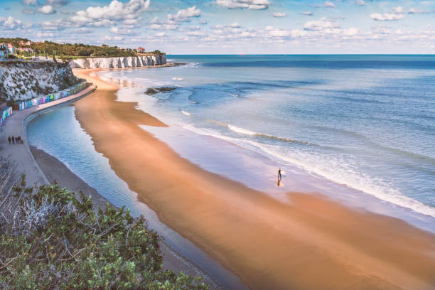 Low tide at Stone Bay, Broadstairs, Kent as summer turns to autumn, a lone surfer walks on the beach and a family on the promenade along side the beach huts and white cliffs. stock photo