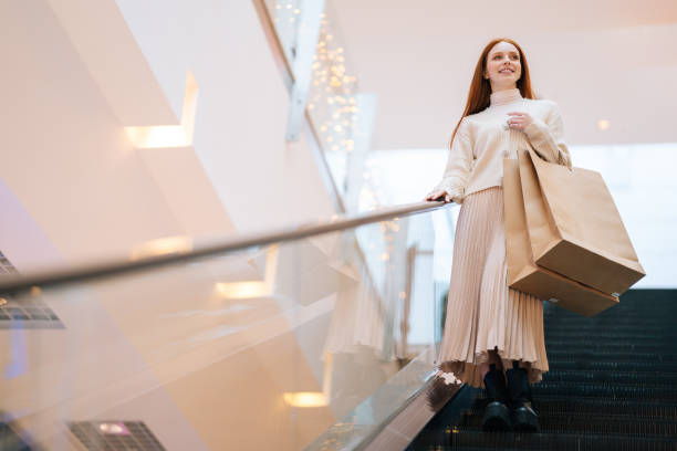 Low-angle view of smiling redhead young woman shopper holding on escalator handrail and riding escalator going down in shopping mall stock photo