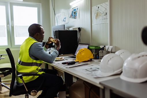 African American man eating a sandwich while working in his office at the construction site