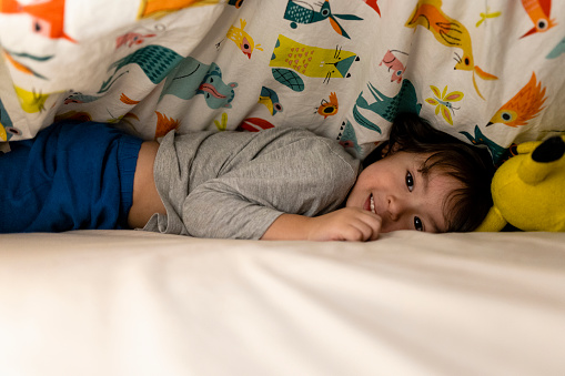 A side-view shot of a young boy wearing pyjamas lying on his front under the duvet in his bed. He is looking at the camera with a cheeky smile, he isn't ready to fall asleep yet.