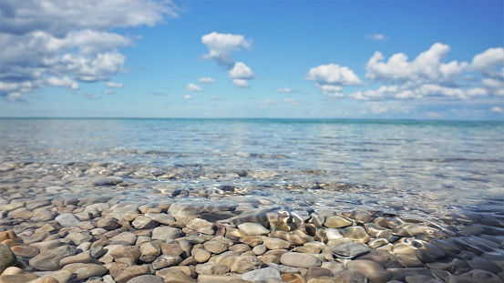 Low angle view of large pebbles in the pristine water Lake Michigan, with puffy clouds in the blue sky above the teal horizon.