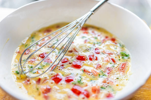 Macro closeup of white bowl plate with balloon whisk cooking omelette with tomatoes, cilantro and red peppers vegetables in homemade kitchen stock photo